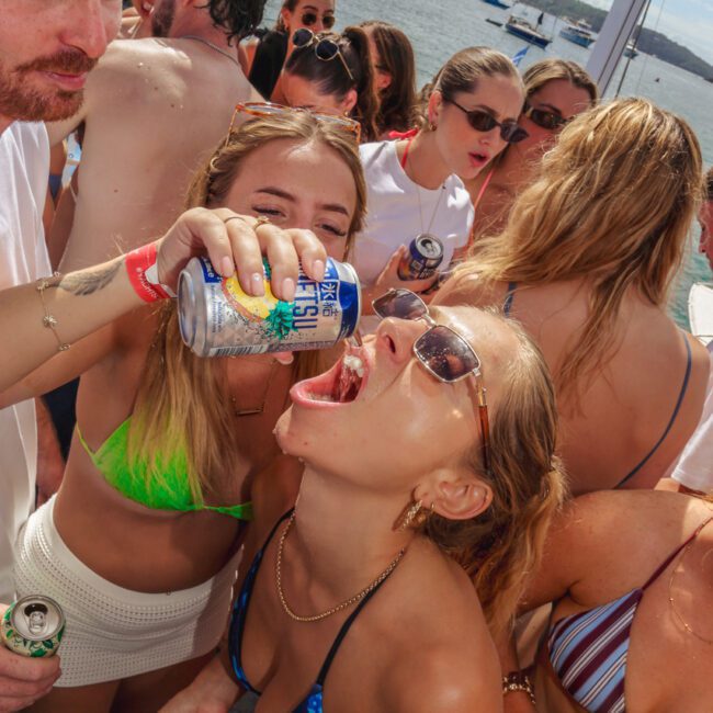 A group of young adults at a lively boat party; one woman in sunglasses and a blue bikini tilts her head back as another pours a drink from a can into her mouth. Others around them chat and hold drinks.