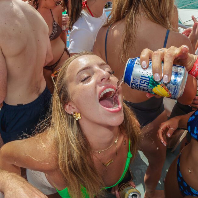 A young woman in a green bikini laughs as another person pours a canned drink into her mouth at a crowded outdoor boat party, surrounded by people in swimwear on a sunny day.