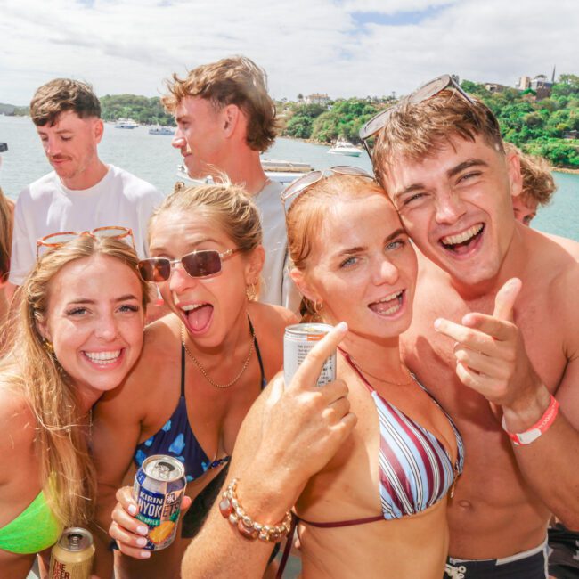 A group of young adults in swimsuits smile and pose together on a boat, holding drinks, with water and greenery in the background on a sunny day.
