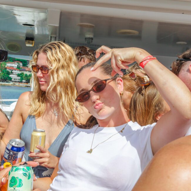 A group of young adults enjoy a sunny day on a boat. One woman in sunglasses strikes a playful pose while holding a drink, surrounded by friends who are also wearing sunglasses and holding cans. The atmosphere is lively and fun.