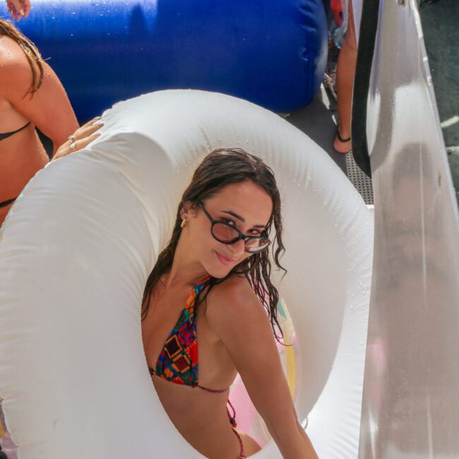 A woman in a colorful bikini and sunglasses smiles while standing inside a white inflatable pool ring at a lively pool or boat party with other people around her.