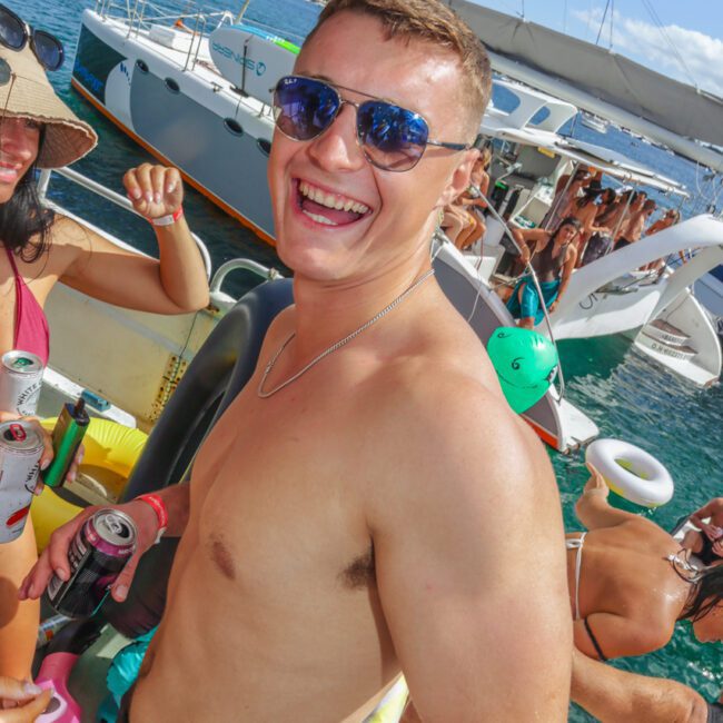 A shirtless man wearing sunglasses smiles at the camera while standing on a yacht during a lively boat party, surrounded by other people, drinks, and water with more boats in the background.