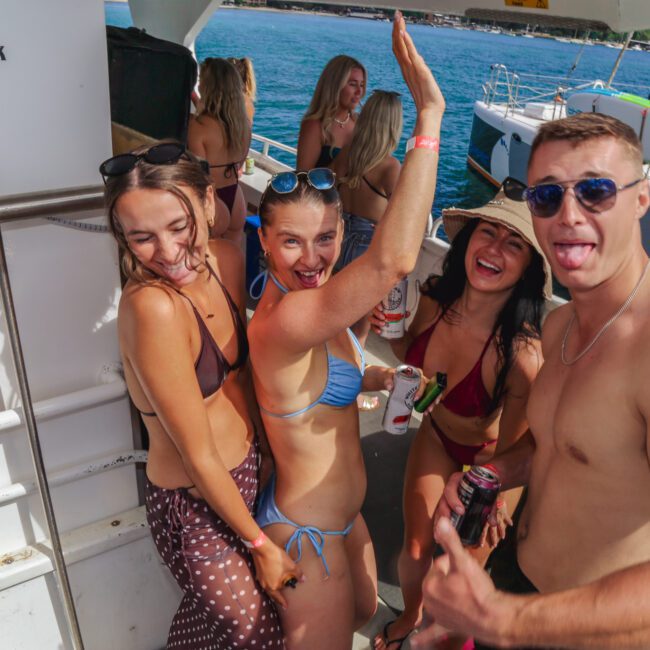 Four young adults in swimwear smile, laugh, and pose for the camera on a boat during a sunny day, holding drinks, with water and other boats visible in the background.