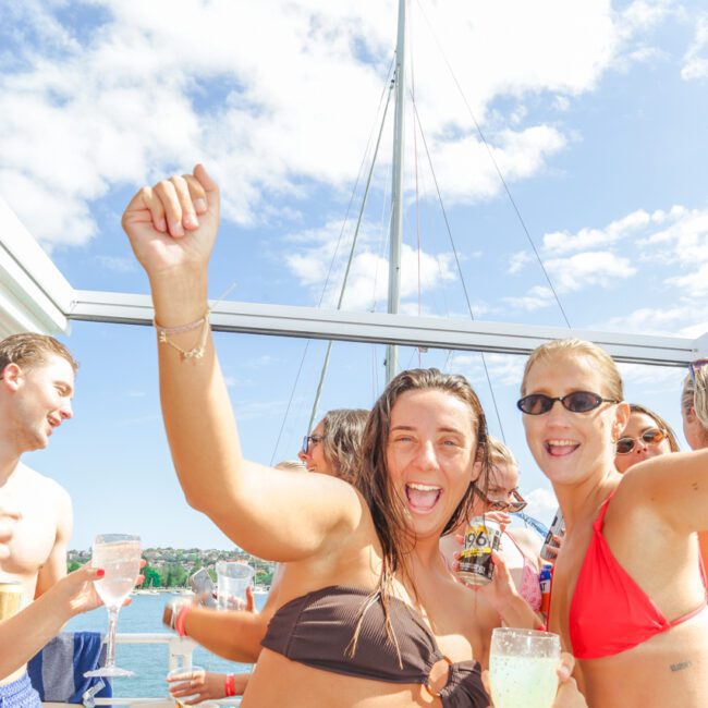 A group of people celebrate on a boat under a sunny sky. A woman in the foreground smiles and raises her arm excitedly while holding a drink. Others around her also enjoy drinks and wear swimwear and sunglasses.