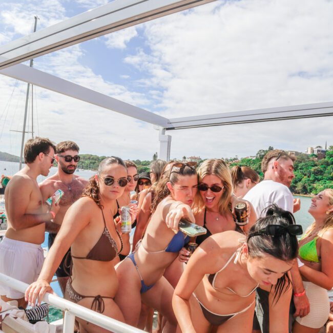 A group of young adults in swimsuits gather on a boat under sunny skies, enjoying drinks and socializing near a railing with water and trees visible in the background.