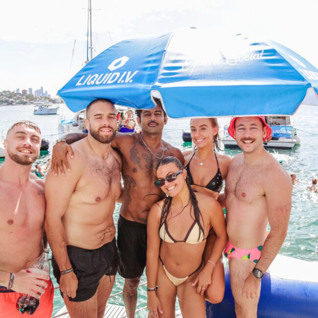 Six people in swimwear pose and smile together on a dock by the water, surrounded by boats and blue umbrellas on a sunny day. The background features clear water and a city skyline.