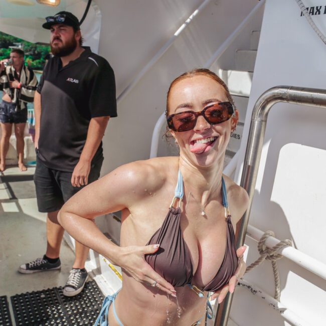 A woman in a bikini and sunglasses playfully poses on a boat, sticking out her tongue and holding her chest. Water drips from her as people stand in the background.