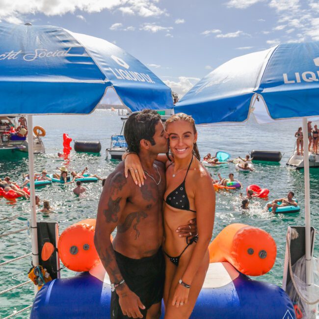 A man kisses a smiling woman on the cheek as they stand under blue umbrellas on a dock. Both wear swimsuits. Behind them, people swim and relax on floats in the water, enjoying a sunny day.