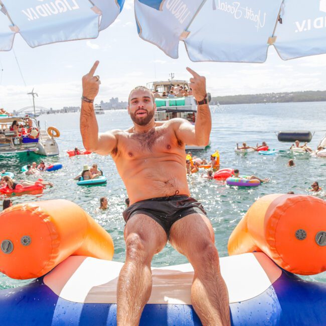 A shirtless man sits on an inflatable water slide, raising both middle fingers. People float in the water on inflatables. Boats and umbrellas are in the background on a sunny day.