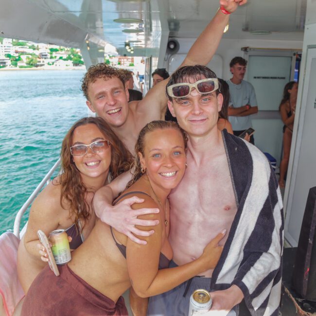 Four young adults in swimsuits smile and pose together on a boat. Two wear sunglasses, and one has a towel around his shoulders. The background shows water, a shoreline with buildings, and other people on the boat.