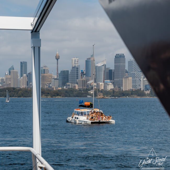 A catamaran with people on board floats on the water, with the Sydney city skyline and Sydney Tower in the background. The photo is taken from another boat, with railings visible in the foreground.