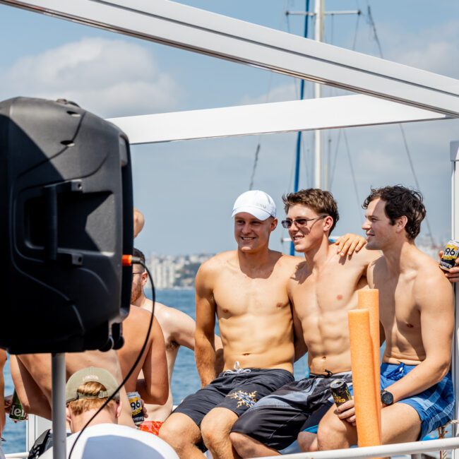 A group of young men in swim trunks sit and stand close together on a boat, smiling and enjoying drinks under sunny skies, with water and city buildings visible in the background.