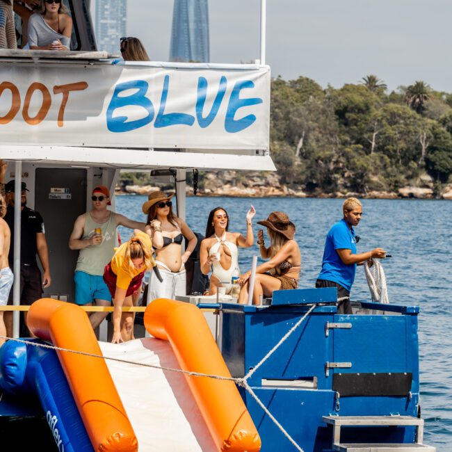 A group of people in swimsuits enjoy a sunny day on a boat with an orange and blue slide into the water. Some are dancing, others are relaxing, and a man stands near the boat's edge. Trees and city buildings are visible in the background.