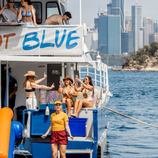 A group of people in swimsuits and summer clothes enjoy a boat party, some sitting and cheering with drinks. The boat is on the water with a city skyline and tall buildings in the background.
