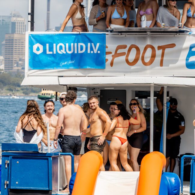 A group of young people in swimwear enjoy a lively party on a boat with a Liquid I.V. banner, docked on the water with a city skyline and landmark tower visible in the background.