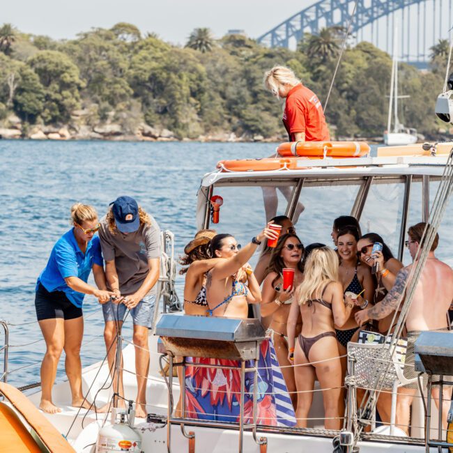 A group of people in swimsuits and casual clothes enjoy a party on a yacht, holding drinks and socializing. The yacht is on the water with greenery and a bridge visible in the background.