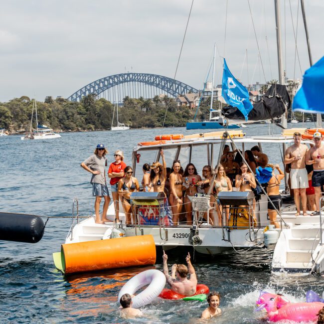 A group of people on a boat enjoy a party, some posing and others swimming with inflatables. The background features water, trees, and Sydney Harbour Bridge under a partly cloudy sky.