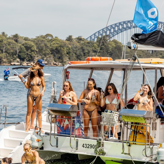 A group of women in swimsuits stand on a boat, holding drinks, while several people relax on floaties in the water nearby. The Sydney Harbour Bridge and trees are visible in the background under a partly cloudy sky.