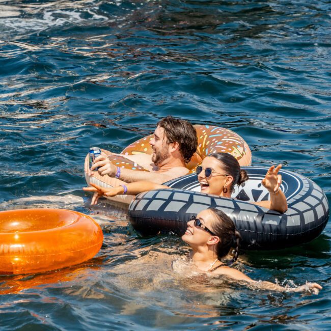 Three people smile and relax on inflatable pool rings in the water, enjoying a sunny day. One person nearby swims, and boats are visible in the background. The Yacht Social Club logo appears at the bottom right.