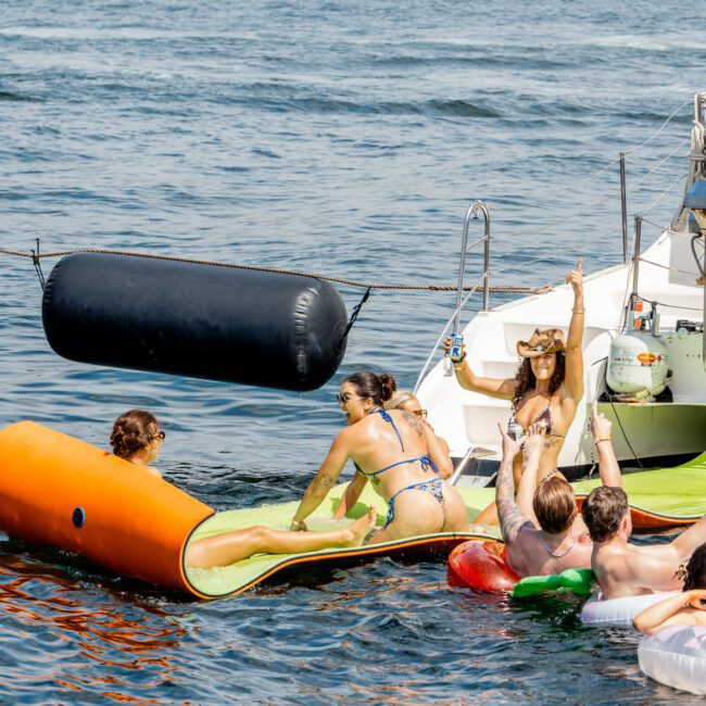 A group of people enjoy a sunny day on the water near a boat, lounging on inflatable pool floats and swimming, with trees visible in the background.