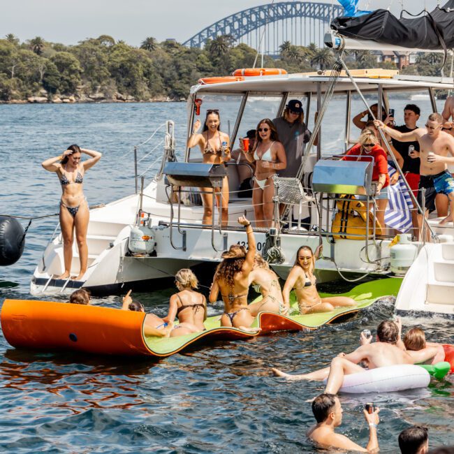 A group of people party on a boat and float in the water nearby; some stand on the deck, others relax on an inflatable, with Sydney Harbour Bridge visible in the background.