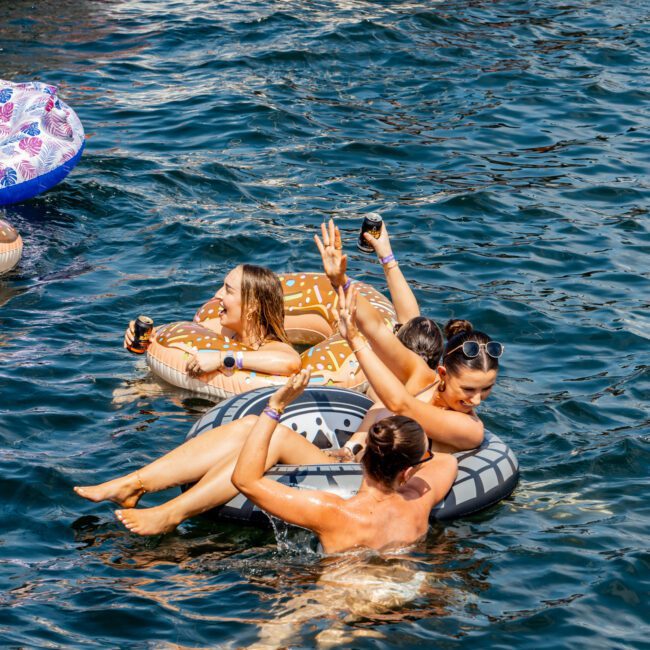 Four people relax on inflatable pool floats in the water, holding drinks and smiling. They are near boats on a sunny day, enjoying a lively atmosphere at a social event.