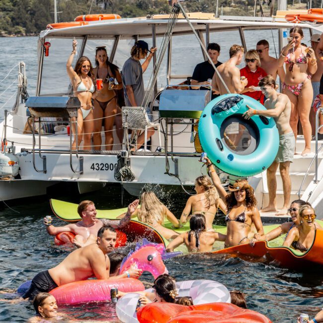 A group of people enjoys a lively party on a boat and in the water, floating on colorful inflatables with the Sydney Harbour Bridge visible in the background. The scene is sunny and festive.