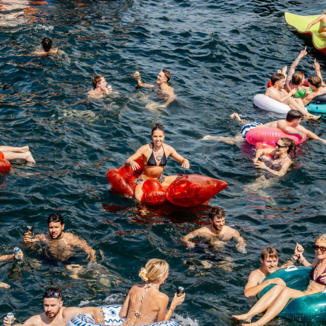 People are swimming and relaxing on colorful inflatables in the sea near a dock, holding drinks and enjoying a lively social event. The atmosphere is festive and energetic, with many smiling faces.