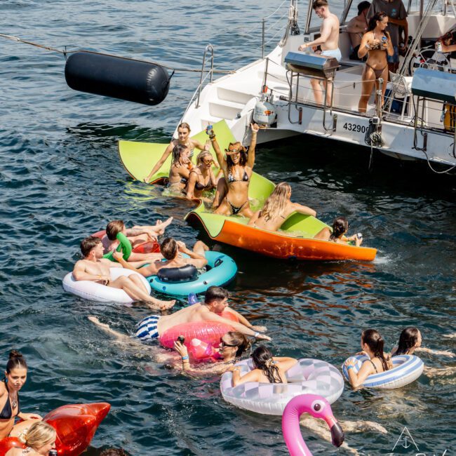 A group of people in swimsuits float on inflatables and paddle boards near a white yacht, enjoying a sunny day on the water. Some are on the boat, while others swim or relax on colorful floats, including a large pink flamingo.