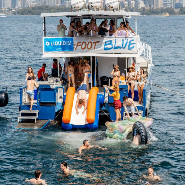 A group of people enjoy a party on a boat with a waterslide, jumping and swimming in the water. The city skyline is visible in the background under a partly cloudy sky.