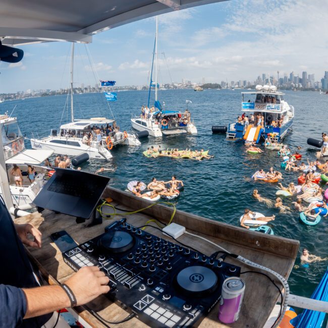 A DJ wearing sunglasses and headphones plays music on a yacht deck, overlooking a lively party with people swimming, floating on inflatables, and relaxing on nearby boats in a sunny harbor with a city skyline in the distance.