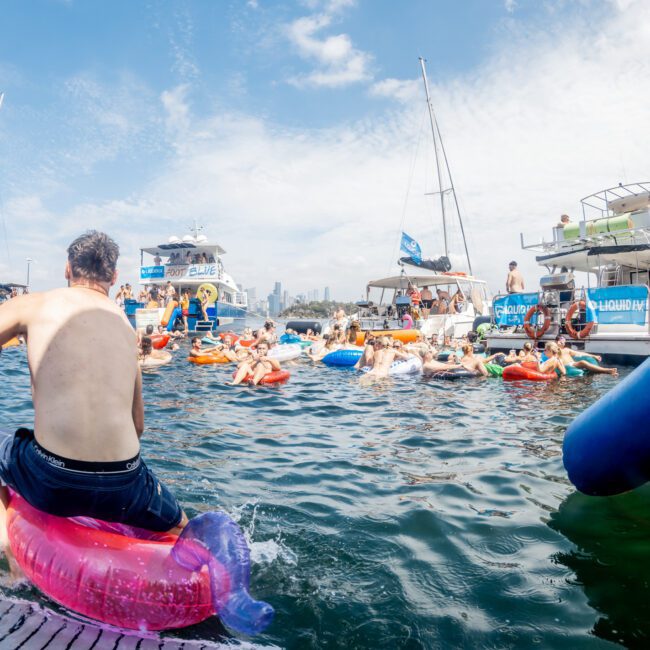 A lively scene on the water shows people on colorful pool floats near yachts under a bright sky, with some swimming and others socializing at a festive boat party.