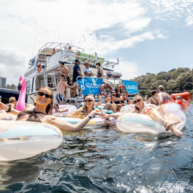 A group of people relax on inflatable pool floats in the water near a large boat. Others are on the boat, socializing under sunny skies. The scene is festive, with bright swimsuits, sunglasses, and a pink flamingo float.