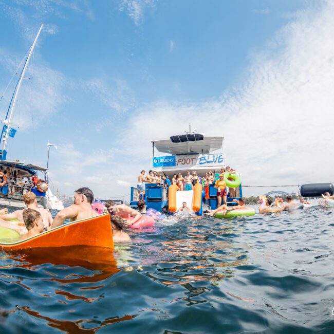 A group of people enjoy a lively party in the sea, floating on colorful inflatables near boats and a floating platform under a partly cloudy sky. Some are swimming, others relaxing, with music and fun in the background.
