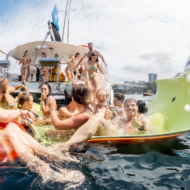 A group of people in swimsuits laugh and relax on colorful floats in the water near a yacht on a sunny day, with others standing and sitting on the boat in the background.