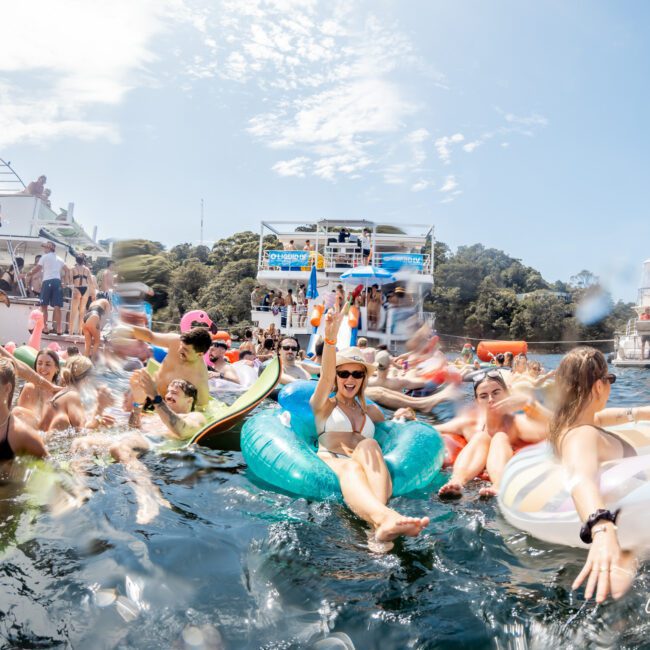 A lively group of people on inflatable floats enjoy a sunny day in the water near boats, laughing, swimming, and socializing, with trees and a partly cloudy sky in the background.