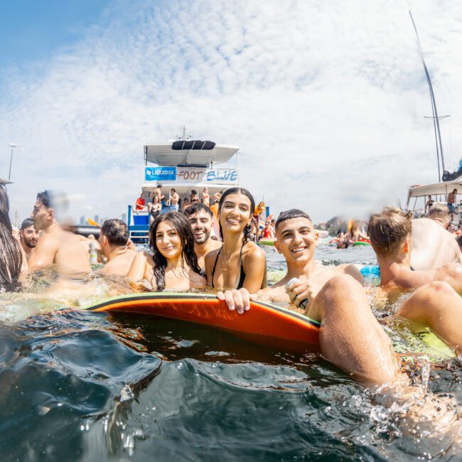 A group of smiling young people in swimsuits gather around a surfboard in the water near boats, enjoying a sunny day at a lively beach or boat party.