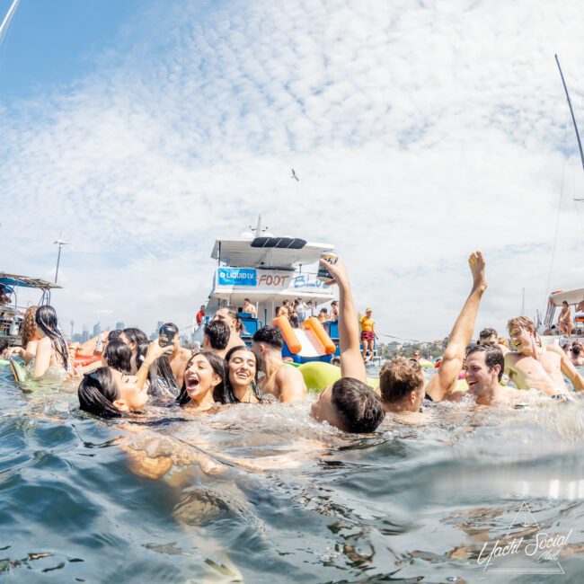 A group of young people laugh and swim together in the water near a floating bar and boats on a sunny day, with blue sky and scattered clouds above.