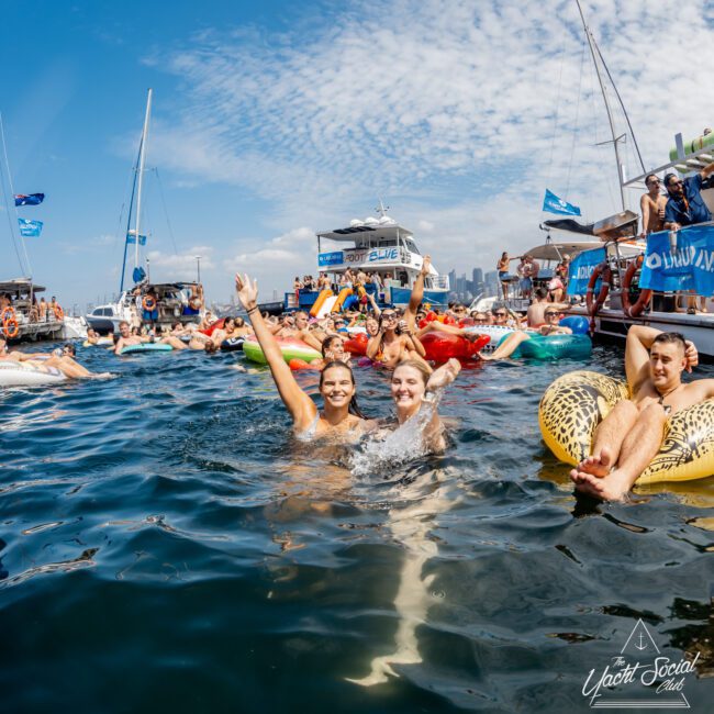People enjoy a lively yacht party on the water, floating on colorful inflatables and swimming near boats under a sunny sky. Two smiling people wave at the camera in the foreground. The city skyline is visible in the distance.