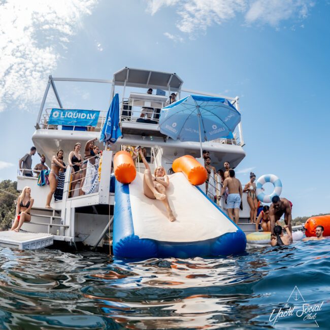A group of people enjoy a sunny day on a double-decker boat with slides, lounging, swimming, and socializing. One person slides down an inflatable slide into the water while others watch or relax nearby.