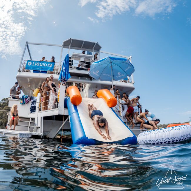 A woman slides down an inflatable water slide from a double-decker party boat into the water, while people watch and relax on the boat and nearby floaties under a blue sky.
