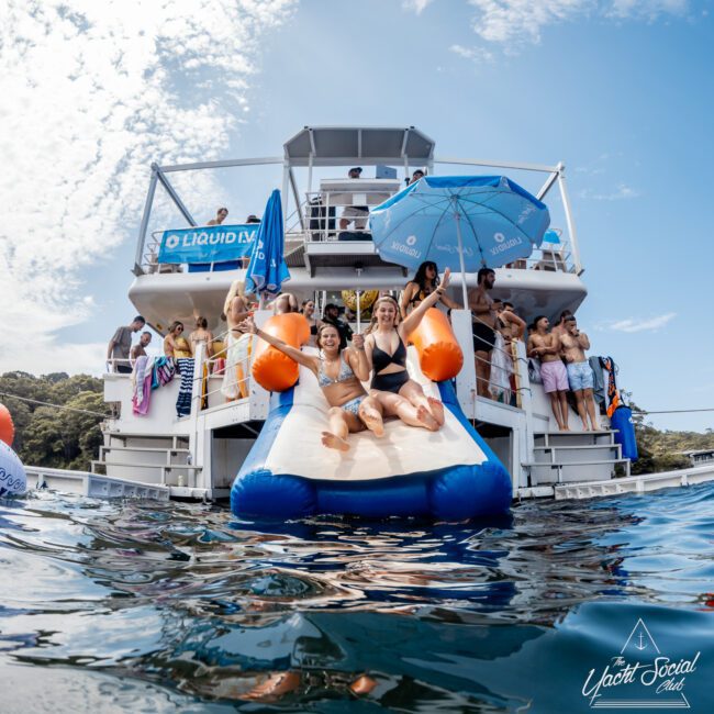 Two women in swimsuits slide down an inflatable water slide from a large yacht into the water, surrounded by people relaxing and socializing on the yacht under blue sky and umbrellas.