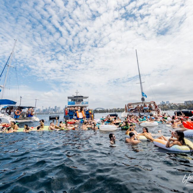 A large group of people in swimsuits float on inflatables and swim in the water between anchored boats during a lively social event on a sunny day, with city buildings visible in the background.