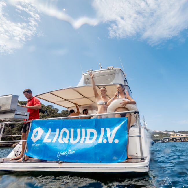A group of people relax on a yacht anchored on the water. Two women in swimsuits stand near the edge, while a “LIQUID I.V.” banner is displayed on the boat. The sky is sunny with scattered clouds.