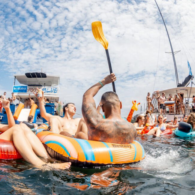 A group of people relax and party on colorful inflatables in the water near boats under a partly cloudy sky, enjoying a sunny day. Some hold drinks while others paddle or talk, creating a lively atmosphere.