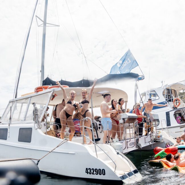 A group of people in swimsuits enjoy themselves on a catamaran, waving and smiling, with others on nearby boats and in the water. The scene is lively and festive under a bright, cloudy sky.