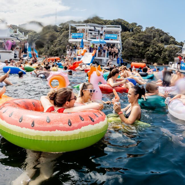 People enjoying a lively pool party on the water with colorful floaties, including a watermelon and unicorn. Boats and a crowd are in the background near green, tree-covered hills. The scene is festive and sunny.