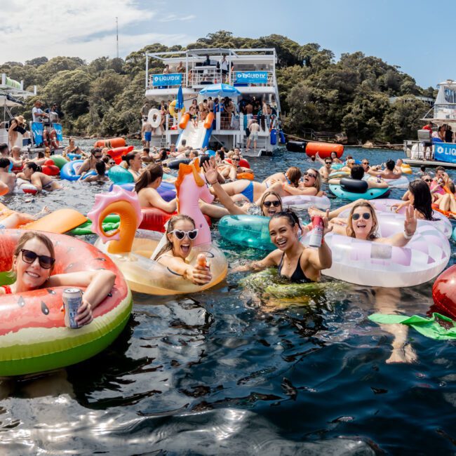 A lively group of people float on colorful inflatable pool toys in the water near docked boats, enjoying a sunny day with drinks and laughter at a festive outdoor party.