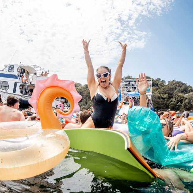 A woman in sunglasses and a black swimsuit cheers with arms raised while sitting on an inflatable float at a crowded boat party on the water. Other people and floats surround her on a sunny day.