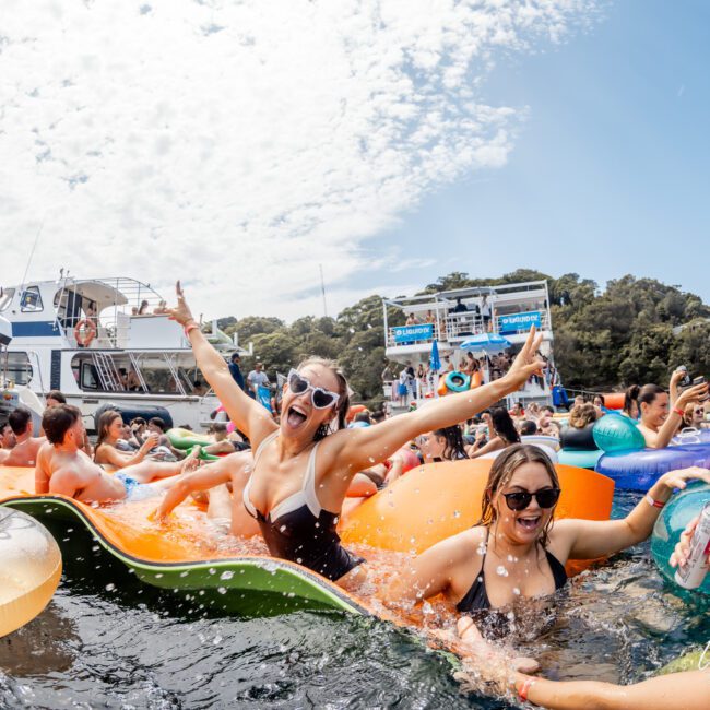A group of people in swimsuits enjoy a lively boat party, floating on colorful inflatables in the water, smiling, laughing, and raising their arms with boats and trees visible in the background under a partly cloudy sky.